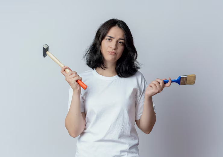 Woman in a white shirt holds a hammer and paintbrush, appearing indecisive. Neutral gray background enhances focus on her expression.