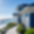 A blue beachfront house with white trim overlooks ocean waves. A curved path and lush greenery surround it under a clear blue sky.