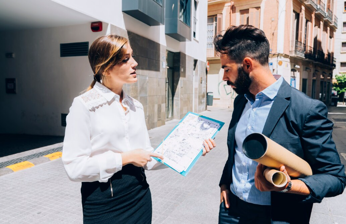 Two people stand on a street, discussing a blueprint. One points at the plan, both in business attire. Urban buildings in the background.