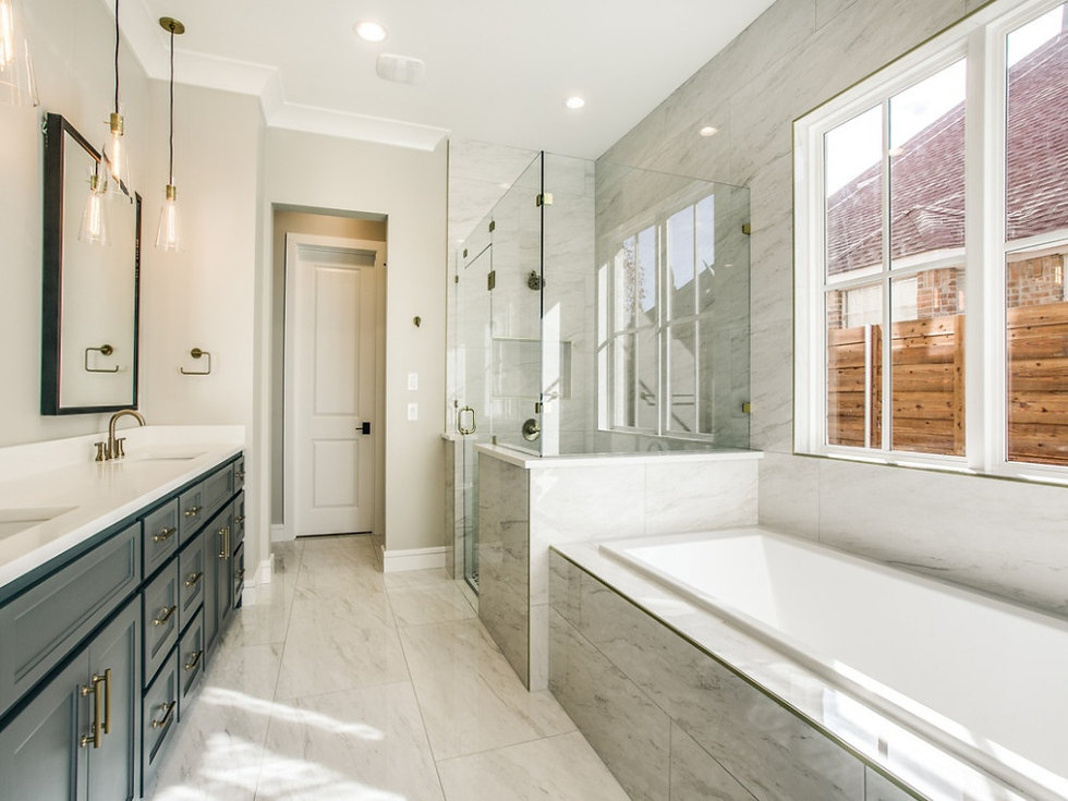 Bright modern bathroom with white marble tiles, soaking tub, glass shower, and dual sinks with blue cabinets. Large window with wood fence view.