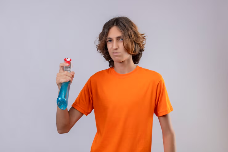 Young person in an orange shirt holding a blue spray bottle with a serious expression. Neutral gray background.