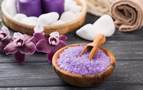 Wooden bowl with purple bath salts and scoop, surrounded by purple orchids, candles, and towels on a rustic gray surface. Relaxing spa ambiance.
