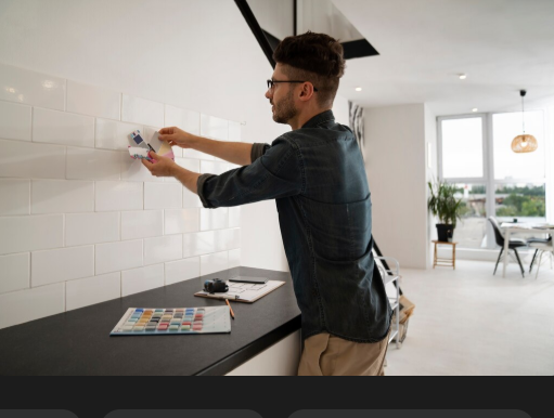 Man in a modern kitchen tests paint colors on a tiled wall, holding a swatch. Bright, airy room with large windows and table.
