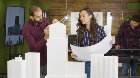 Two people in an office setting discuss a building model, holding blueprints. Another person works at a desk. Background has wood paneling.