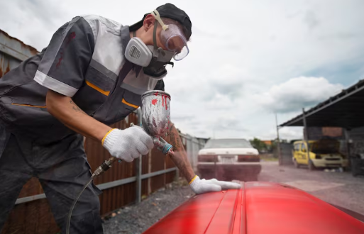 Person in protective gear spray-painting a red car in an outdoor setting. Cloudy sky, parked cars, and a metal fence in the background.