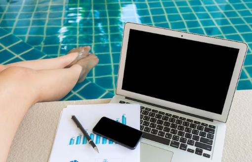 Laptop, phone, and charts on a table by a pool. Legs dangle in water. Blue tile pattern. Relaxed, work-from-pool vibe.