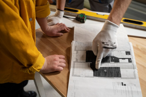 Two people examine a floor plan on a wooden table. One wears a yellow jacket, the other points wearing gloves. A tape measure lies nearby.