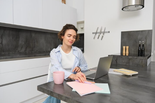 Woman in a modern kitchen using a laptop, holding a pink mug, with notebooks nearby. Gray and white decor creates a calm atmosphere.