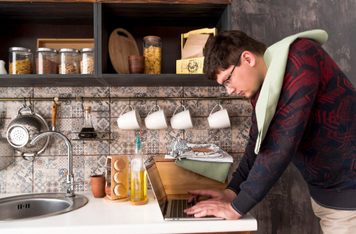Man with a towel over shoulder uses a laptop in a kitchen, surrounded by hanging cups and stored pasta. Patterned tiles add warmth.