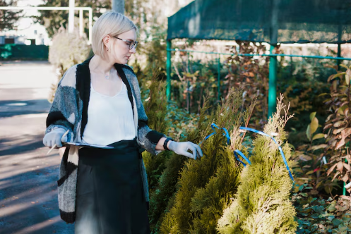 Woman with short hair and glasses examines plants in a greenhouse. She's holding a clipboard, wearing a gray sweater, looking focused.