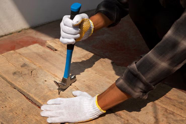 Person wearing white gloves uses a blue-handled hammer on a wooden plank. Sunlit, outdoor setting with red-tiled floor.