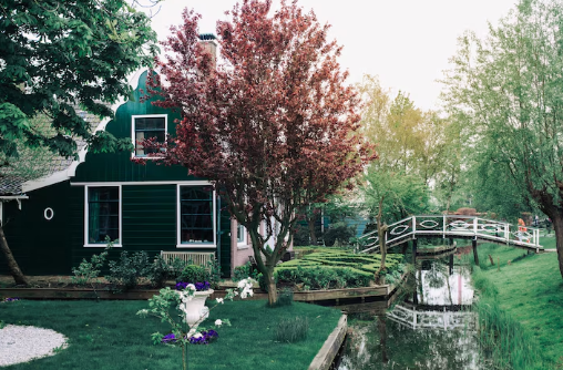Green house with red tree in garden, surrounded by lush greenery. Wooden bridge over narrow canal, serene and picturesque setting.