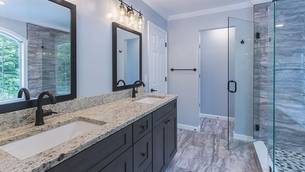 Modern bathroom with dual sinks, granite countertop, dark wood cabinetry, glass shower, gray tiles, and soft lighting. Large window view.