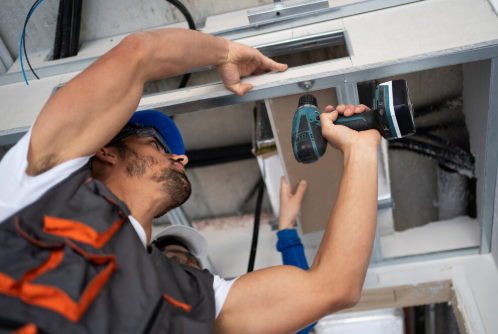 Two construction workers install a ceiling panel. One holds a drill, the other assists. They're focused, wearing safety gear in an industrial setting.
