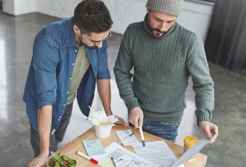 Two men in casual clothes review documents at a table with pens, a salad, and takeout box. One points with a pen. Bright, focused mood.