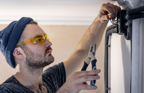 Man wearing a beanie and safety glasses uses pliers to work on electrical wiring. Indoor setting, focused expression, neutral colors.