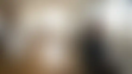 Woman in a grey sweater sits on a beige sofa holding a mug, gazing out a window. Modern living room with wooden floors and a kitchen.