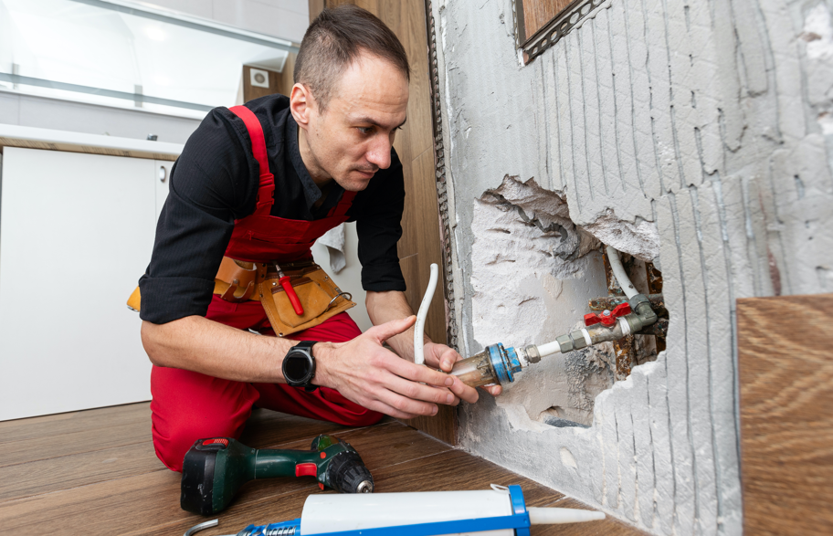 Man in red overalls doing plumbing work, fixing a pipe in a wall hole. Tools like a drill and caulking gun are on the wooden floor.