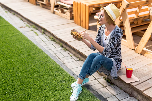 A woman in a hat sits on a curb, eating fries from a box. She's in casual attire with sneakers. Beside her is a red drink cup. Bright, sunny day.