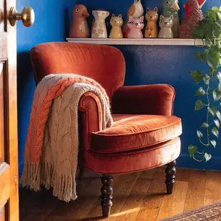 A cozy orange armchair with a draped knit blanket sits on a wooden floor. Shelf with quirky animal vases in a colorful room.