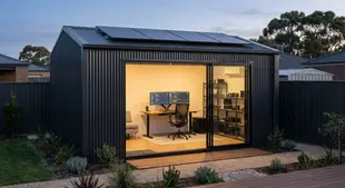 Modern shed office with solar panels. Inside, dual monitors on a desk display code. Warm lighting, minimalist decor, evening setting.