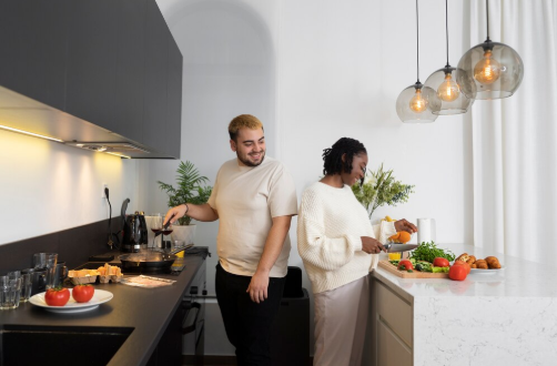 Couple cooking together in a modern kitchen. One is frying, the other chopping vegetables. Warm lighting, white walls, and black cabinets.