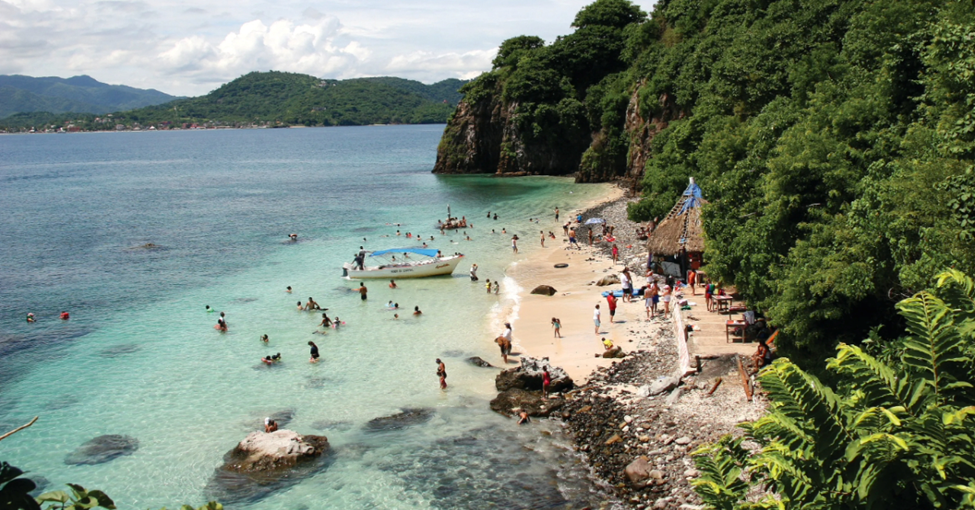 People swimming and relaxing on a rocky beach with clear blue-green water. Lush green hills and a boat are visible. A lively, sunny scene.