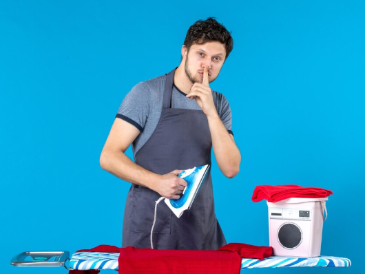 Man ironing red clothes, finger to lips, suggesting silence. He wears a grey apron. Blue background, washer nearby. Playful mood.