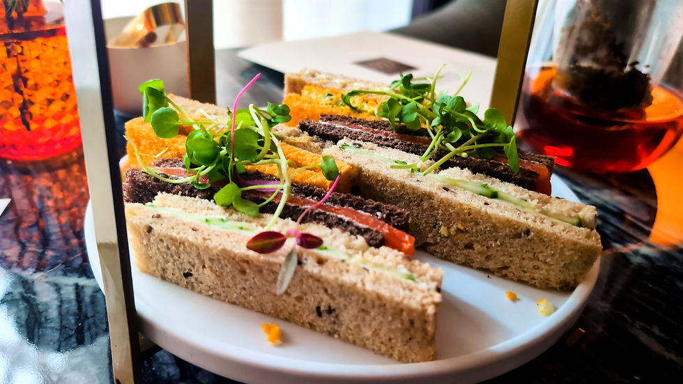 Assorted tea sandwiches topped with microgreens on a white plate beside amber tea glasses in a café setting.