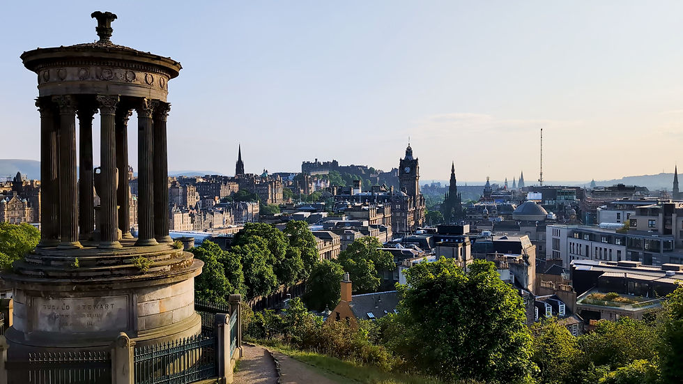 Dugald Stewart Monument overlooks Edinburgh skyline at sunset, with green hills, historic buildings, and clear sky.