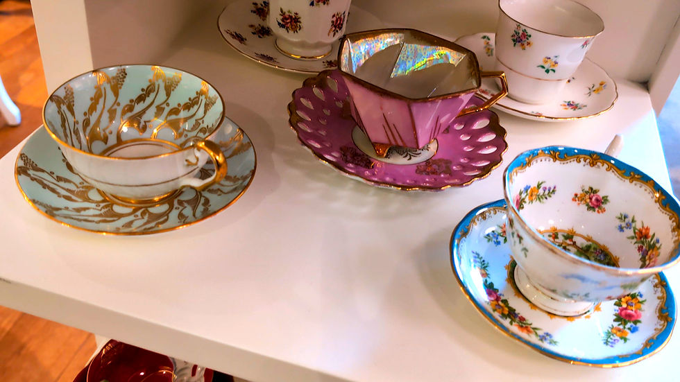 Assorted vintage floral teacups and saucers with gold trim on a white shelf, in white, blue, pink, and purple hues.