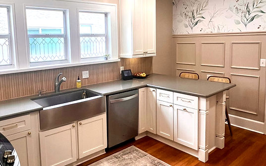 A kitchen with cream shaker cabinets, gray counters, taupe wainscoting, floral wallpaper, wood ribbon tile backsplash, and stainless apron sink.