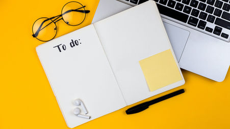 Open to-do list notebook with glasses, laptop, pen, earbuds, and sticky note on a yellow background.