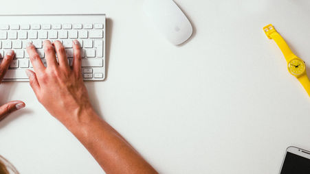 a woman typing on a white keyboard, with an apple mouse and yellow wristwatch beside her