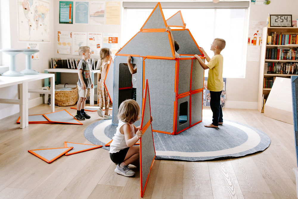 Three children collaboratively build a tall house-shaped structure from Superspace magnetic felt panels in a bright, modern home playroom, with extra panels scattered on the floor mid-build.
