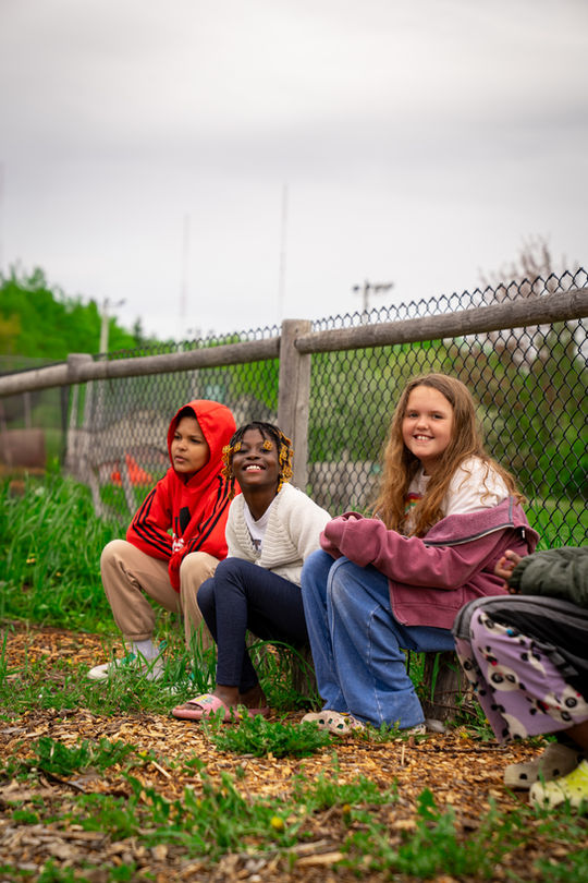 Three young kids smile at a camera in the garden.