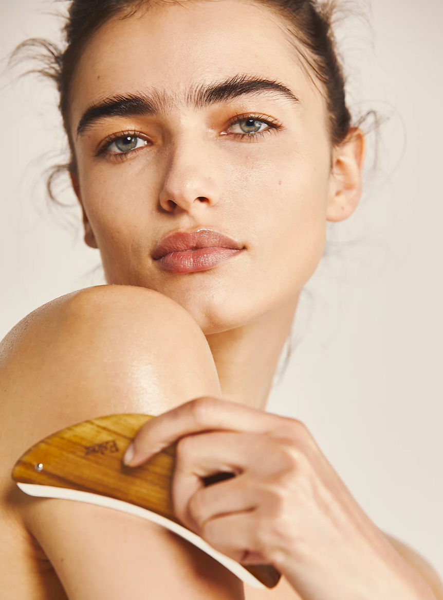 A person uses a wooden gua sha tool on their shoulder. The setting is neutral, with soft lighting highlighting their relaxed expression.