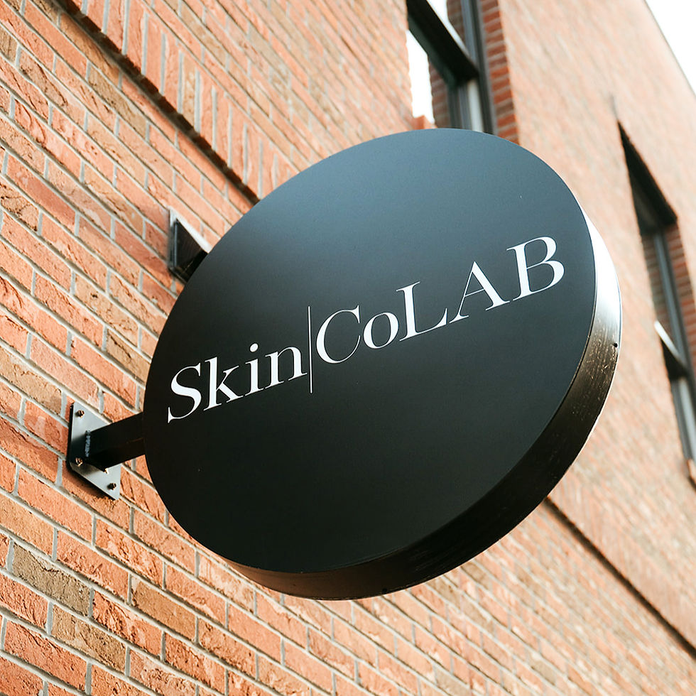 Black oval sign with white text "Skin CoLAB" mounted on a red brick wall, angled view showing building in the background.