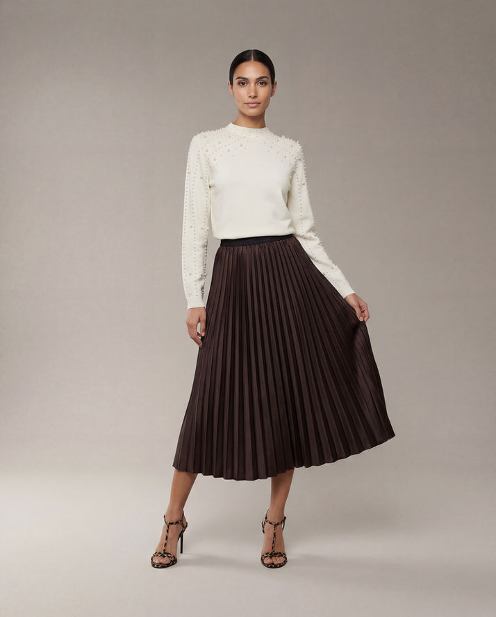 Full-length studio image of a woman in a cream embellished top, holding out her brown pleated skirt.