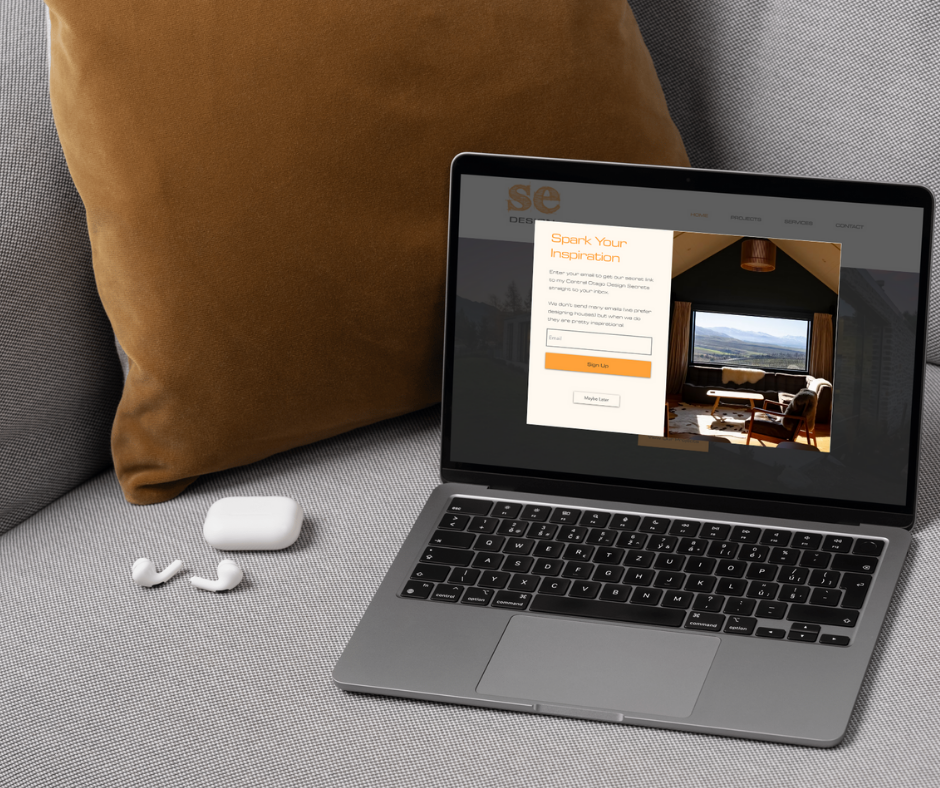 Laptop on gray couch with a brown pillow. Screen shows a website, "Spark Your Inspiration" prompt visible. White earbuds beside it.