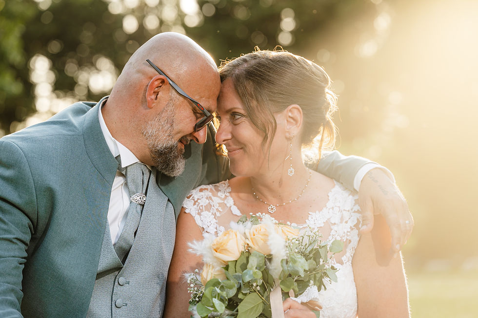 Les mariés se touchent le front et sourient avec tendresse en extérieur ; la mariée tient un bouquet, baignée dans la lumière dorée du soleil couchant.