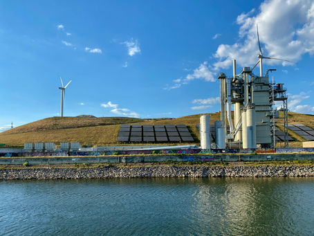 Landscape featuring solar panels on a grassy hill and white wind turbines near a river and industrial facility.