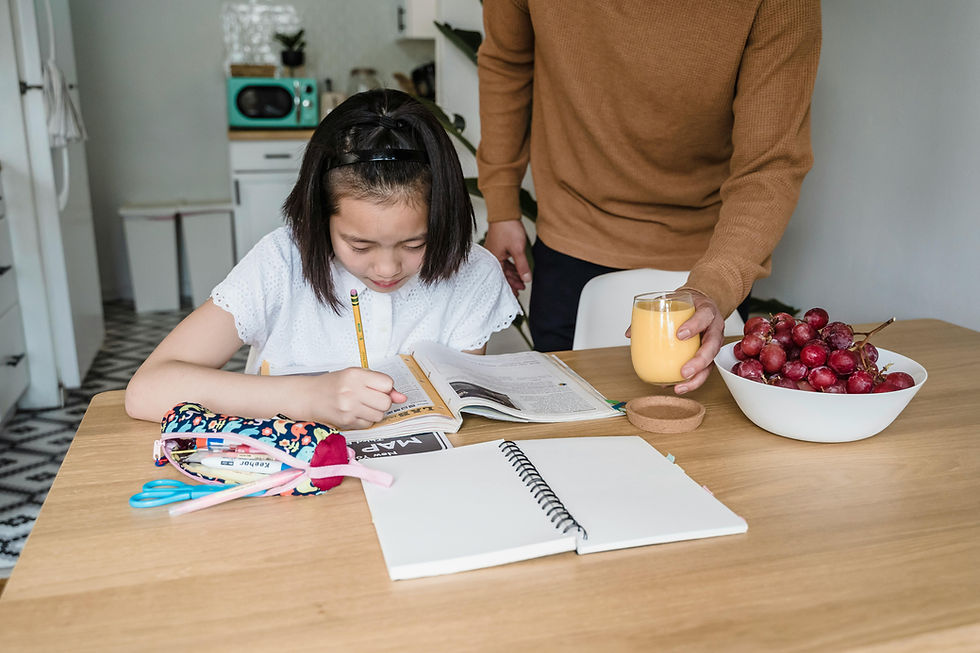 Young girl studying independently at a kitchen table with books and stationery while a parent gently offers a glass of juice, showing a calm home learning routine.