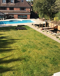 Swimming pool, paved terrace and lawn beside cedar decking and a pond bridge.