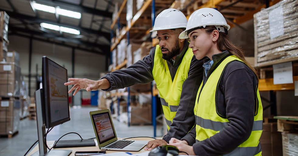 Dois trabalhadores de capacete e colete refletivo analisam um monitor num armazém; ele aponta para a tela.