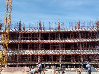 Prédio em construção com múltiplos pavimentos, inteiramente envolvido por andaime fachadeiro metálico vermelho, e trabalhadores atuando no térreo.