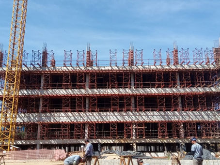 Prédio em construção com múltiplos pavimentos, inteiramente envolvido por andaime fachadeiro metálico vermelho, e trabalhadores atuando no térreo.