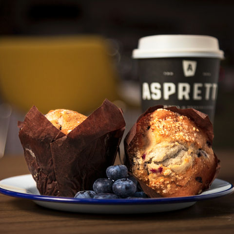 Two blueberry muffins on a white plate with a blue rim, next to a cup of coffee on a wooden table. The muffins are wrapped in brown paper and the coffee cup has the Aspretto logo on it