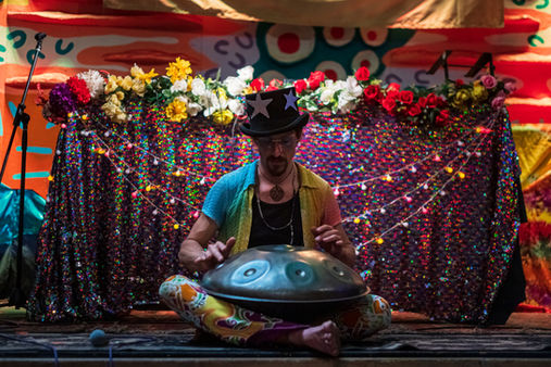 Musician playing a handpan on stage in a colourful festival setting.