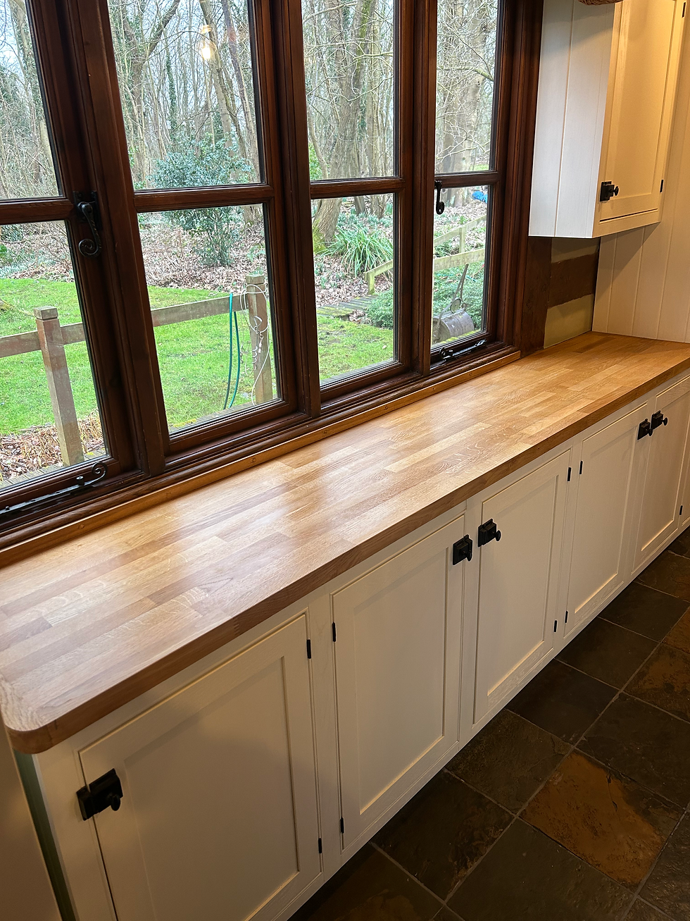 Kitchen window seat with wood countertop and white cabinets, overlooking a wooded yard through large brown-framed windows.